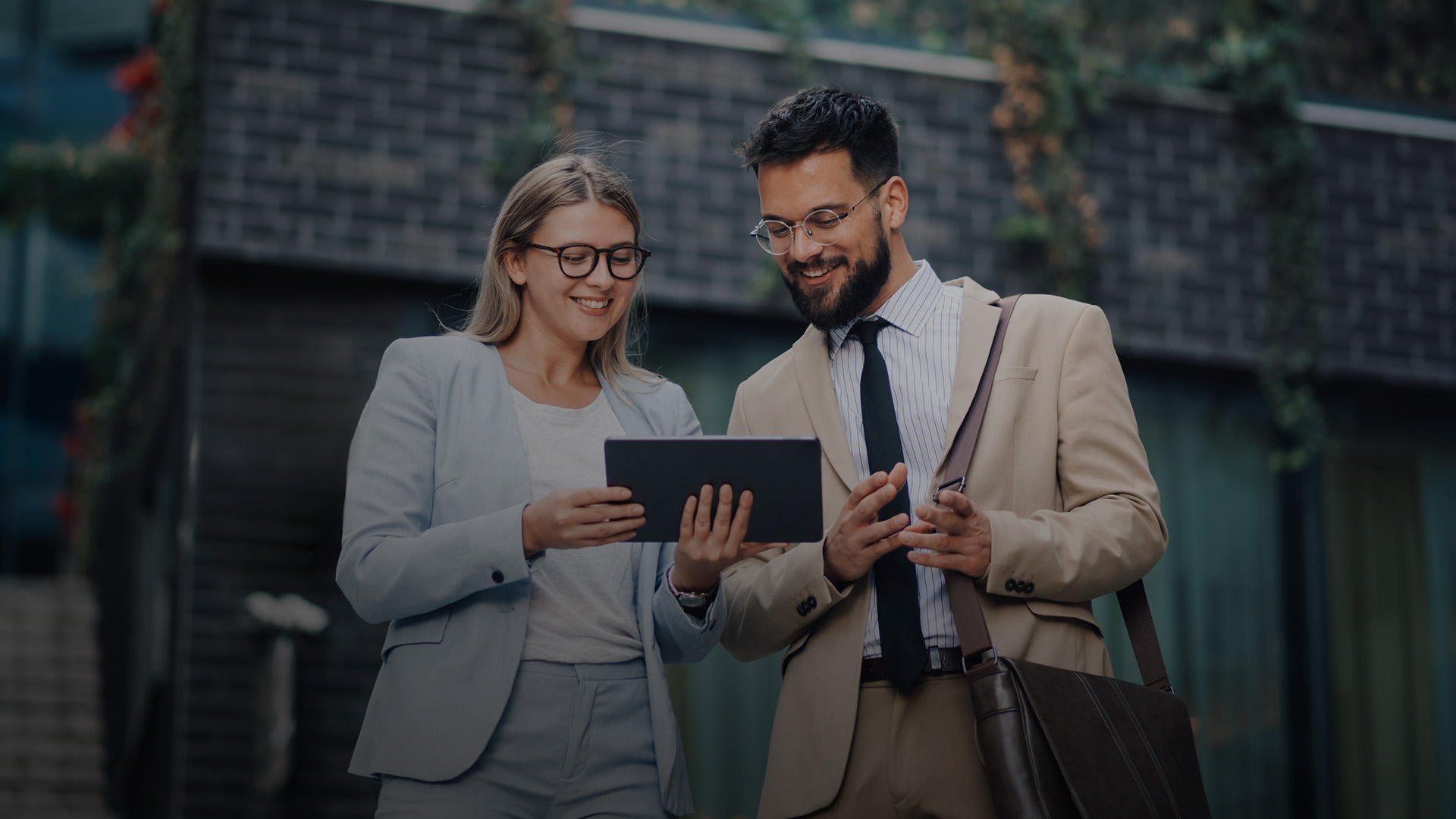   Two business professionals standing outdoors, reviewing information on a tablet. Both are smiling and dressed in formal attire.