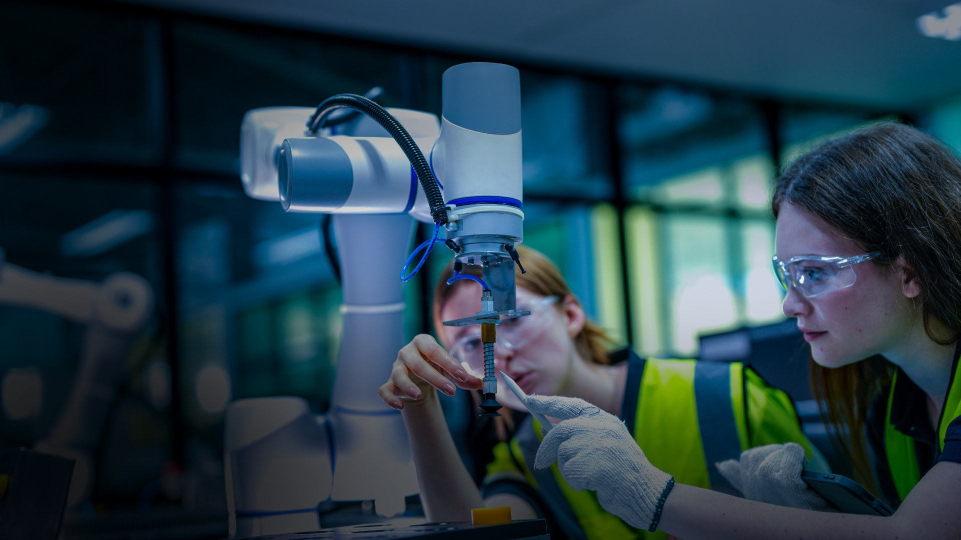 Two people wearing safety vests and protective goggles carefully operate or inspect a robotic arm in a lab or industrial workspace, focusing on a small component held by the robot’s tool.