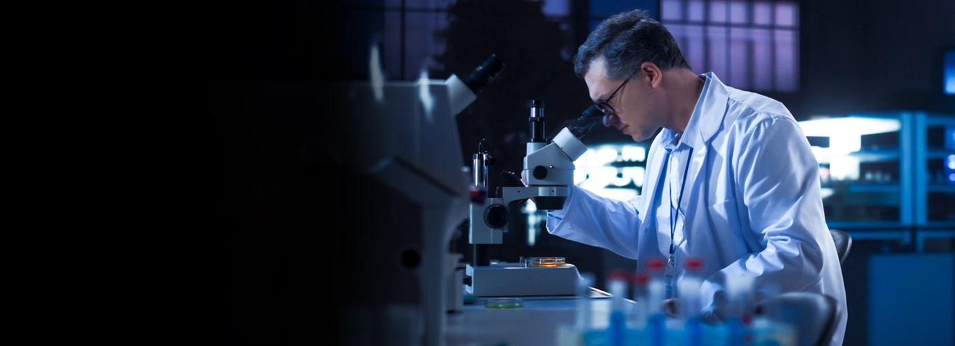 A scientist in a white lab coat looks through a microscope in a modern laboratory, with scientific instruments, sample containers, and illuminated equipment visible in the background.
