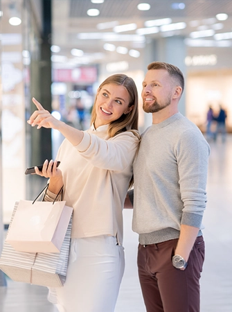 Two people walking inside a bright, modern shopping mall, smiling and carrying multiple shopping bags while one of them points ahead.