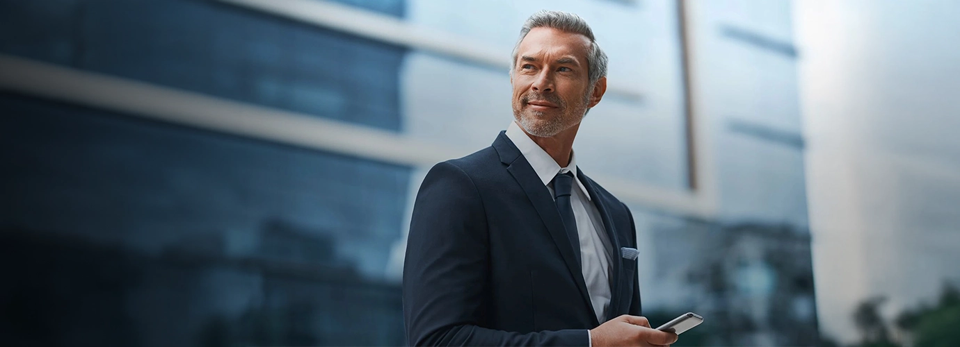 A business executive in a navy suit holding a smartphone outside a modern workplace, representing application managed services, leadership, and enterprise digital operations.