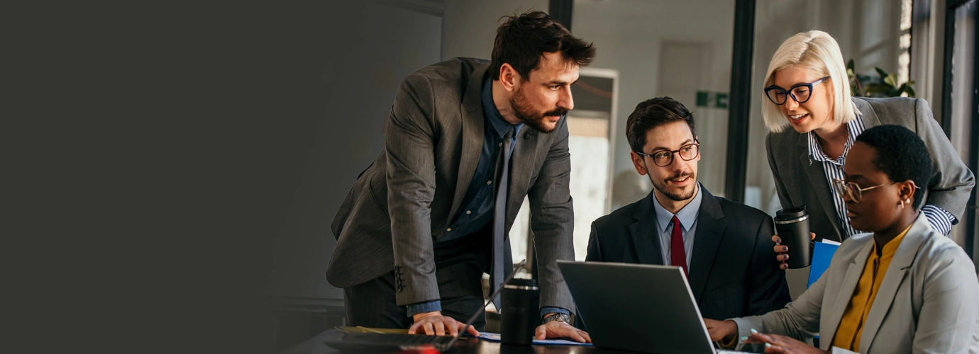 Four business professionals collaborate around a laptop in a modern office, discussing ideas and reviewing information during a team meeting.