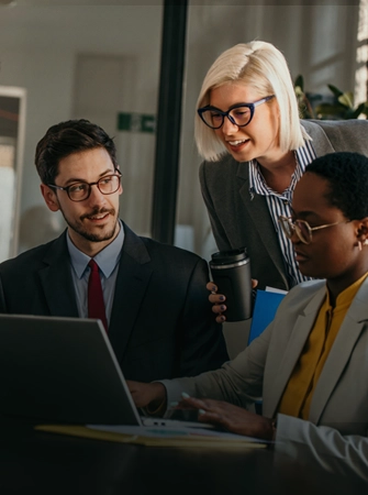 Four business professionals collaborate around a laptop in a modern office, discussing ideas and reviewing information during a team meeting.